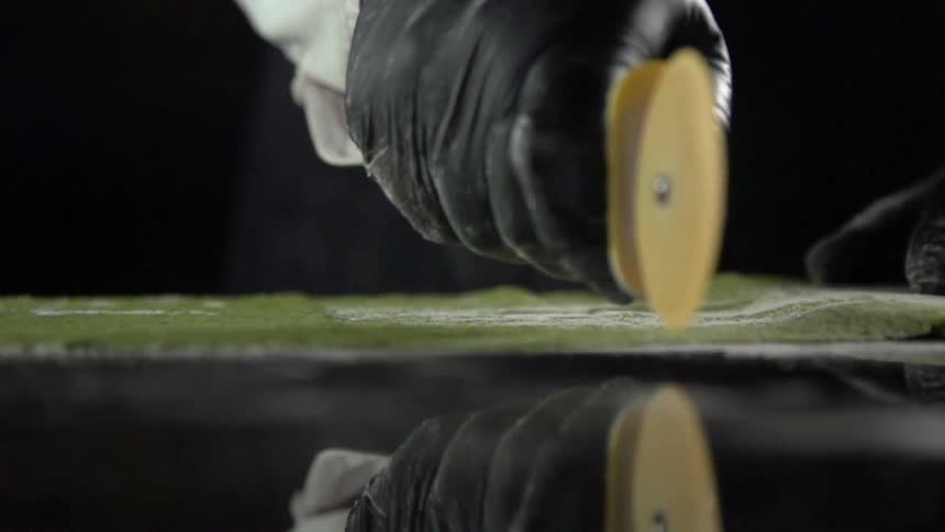 A person wearing black gloves uses a pizza butter to slice green dough