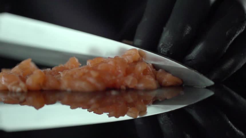 Fresh raw salmon is sliced by a gloved hand on a mirrored surface with a sharp knife
