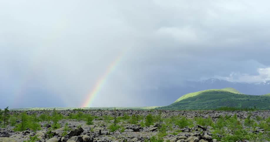 Rainbow occupies horizon as shadow of clouds flow over low green mountain near lava fields