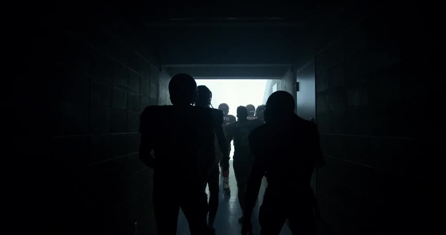 A football team walks through the tunnel towards the stadium holding up their helmets and football in excitement