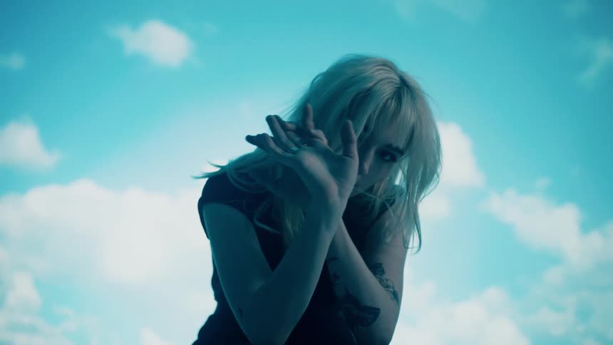 A woman poses with her hands in front of her face as a digital sky plays behind her
