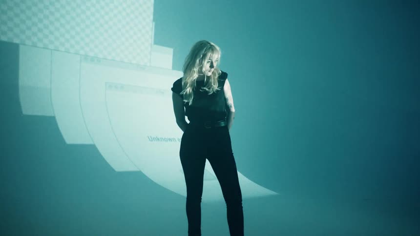 A white woman dressed in black stands on a stage with a giant projection screen lit up behind her