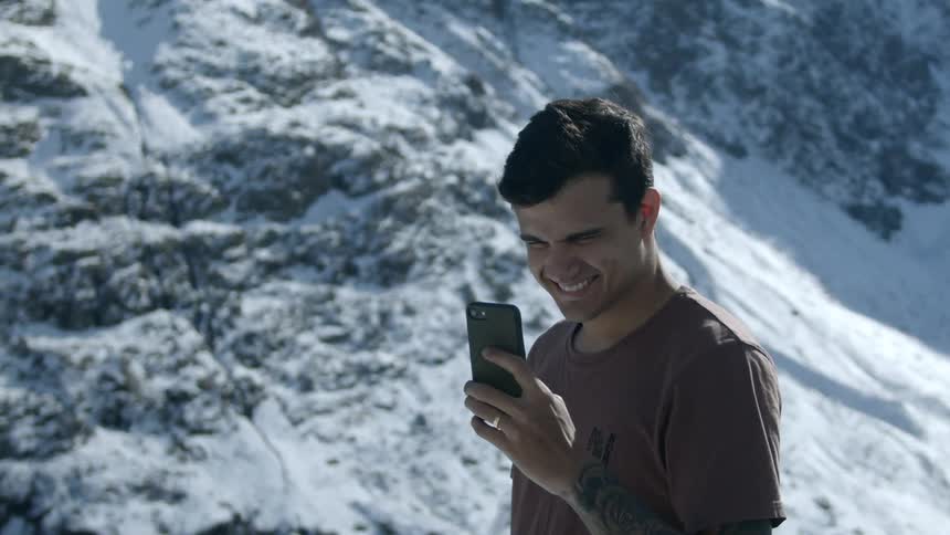A white man stands and smiles into a smartphone as a mountain covered in snow looms behind him