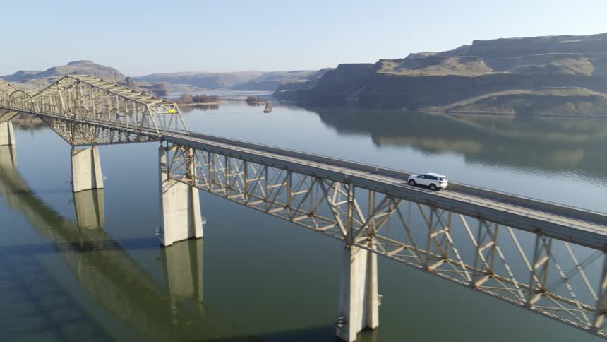Dynamic ascent following a white SUV as it starts to cross a bridge surrounded by hills and rocky cliffs