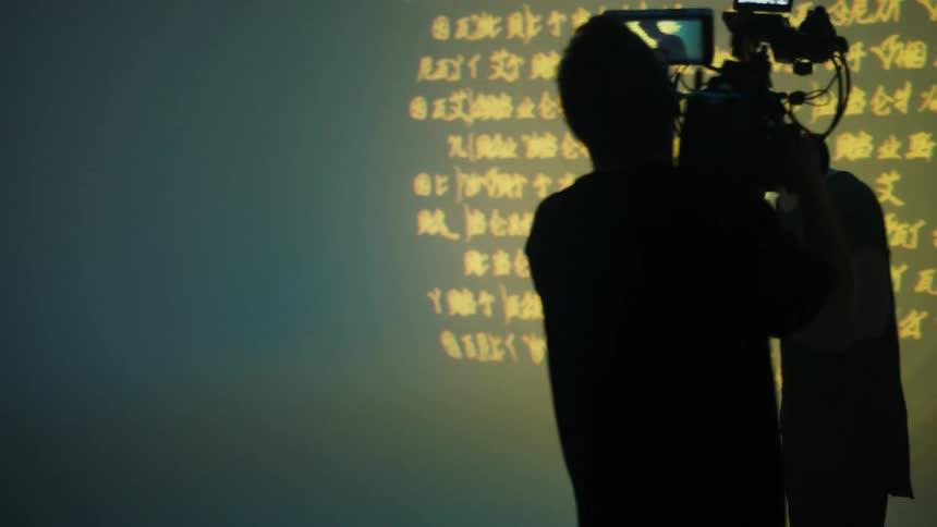 A cameraman films another man working on a laptop that is projecting text onto a giant screen
