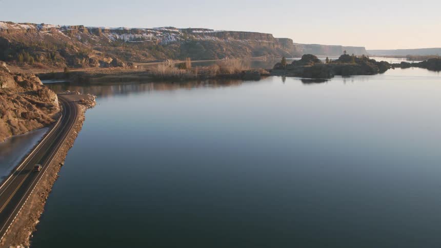 A car drives on a two lane road along the rugged shoreline of a glass calm lake