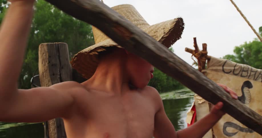 A young boy uses a log to paddle a small handmade boat in a lake