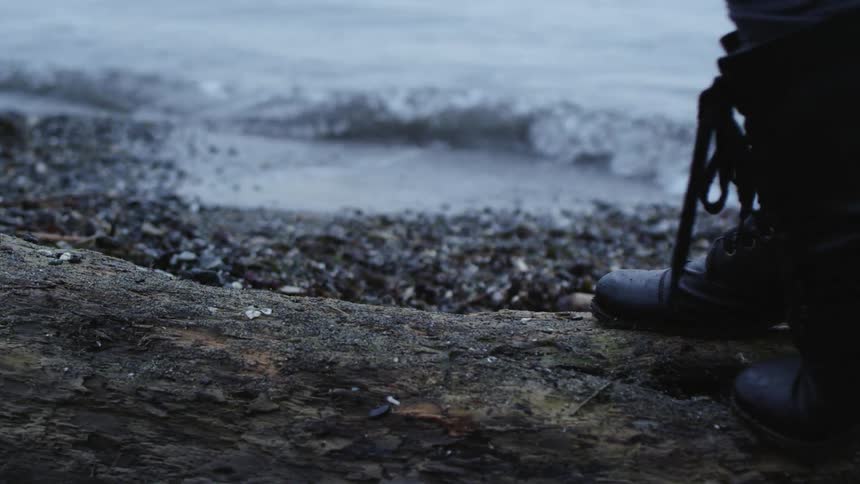Female wearing laced black ankle boots carefully walks across a piece of driftwood on a coast of the beach as the ocean water washes up on the debris in the sand