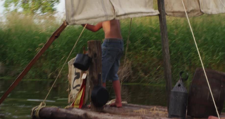 Two boys sail down a river on a handmade raft