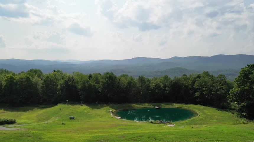 A pond with a small wooden dock in a walled off grassy field with mountains behind