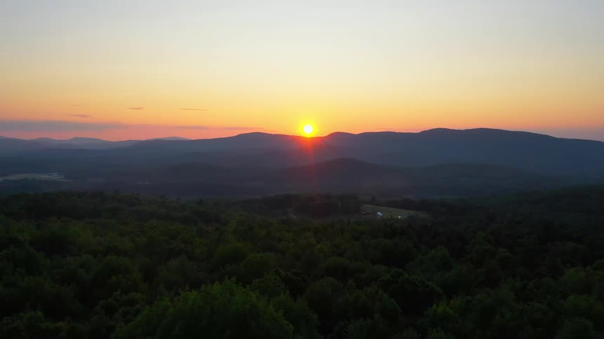 Sun hovers over mountains near forest turning the sky orange