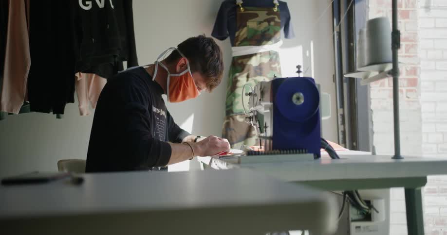 A man sits in a clothing store and sews a mask
