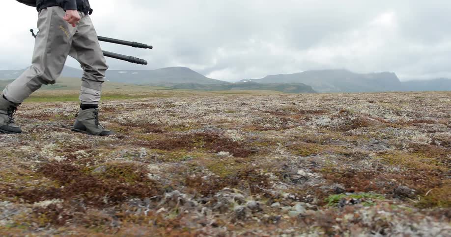 A man is walking over a field holding a tripod on a cloudy day