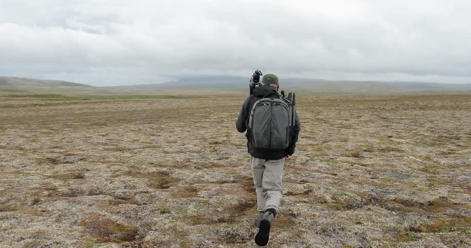 A man carrying a tripod and backpack walks across a plain