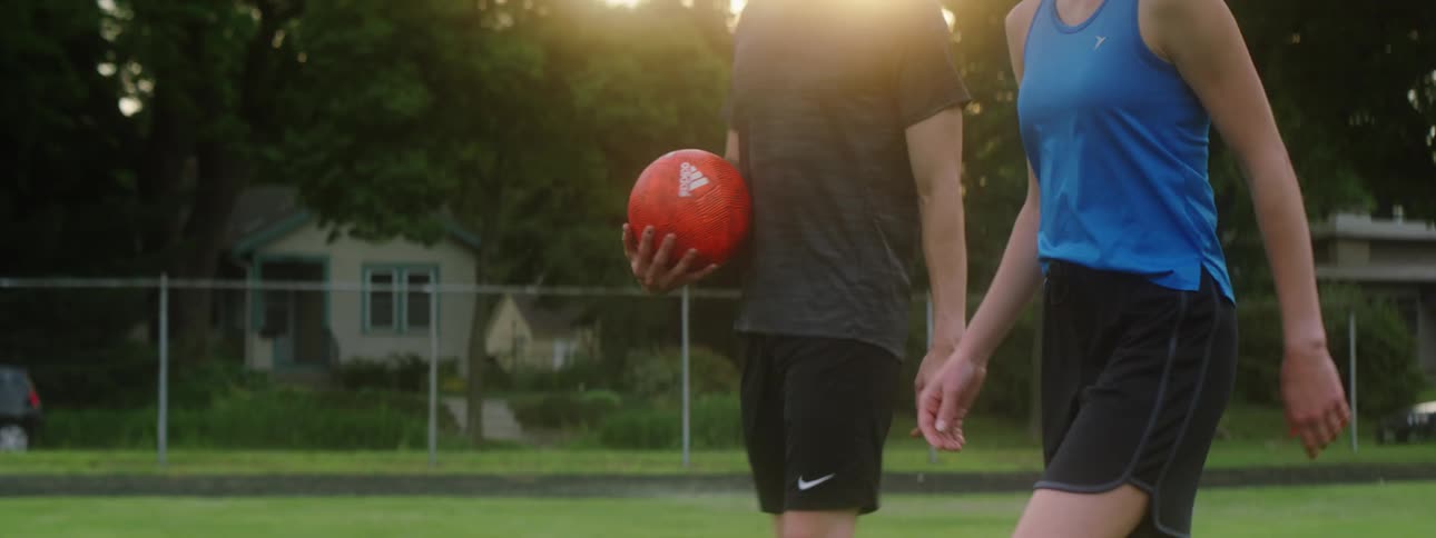 A girl and boy walk along and then drop a ball and kick it