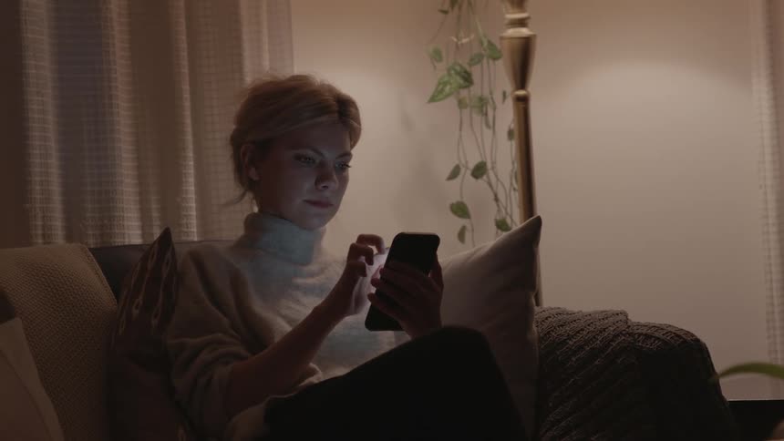 A white woman sits on a couch with her smartphone and pets her cat
