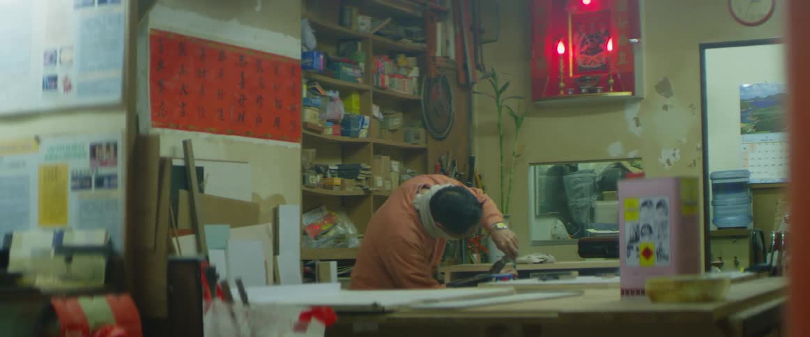A white man works on a piece of wood in a cluttered room