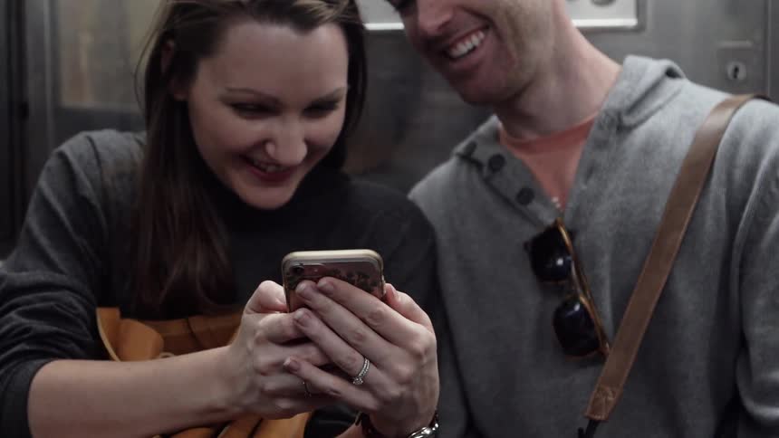A man and a woman laugh over a smartphone while on a subway car
