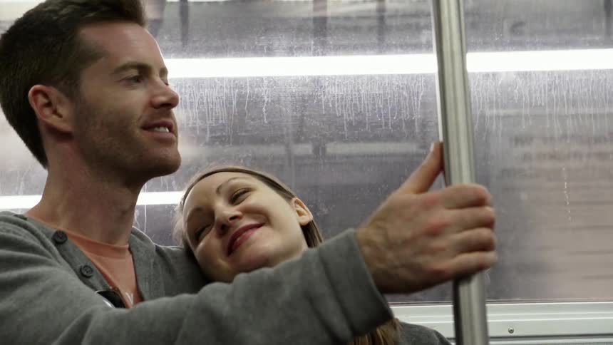 A woman gazes up and smiles and leans back against a man while they are both sitting on a moving subway train
