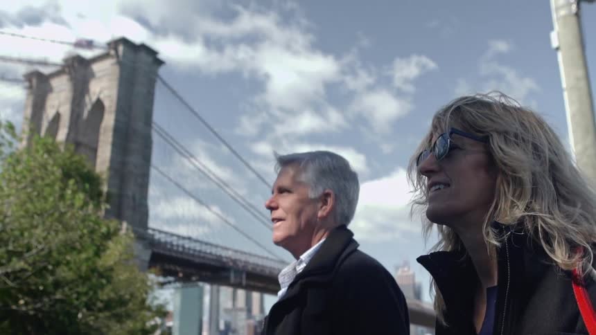 A man and woman walking a city street with a bridge and buildings in the background