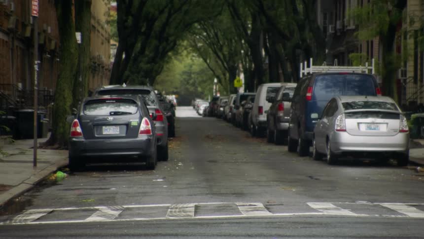 A woman crosses the street in a crosswalk in front of several cars parked in front of apartment buildings in a city