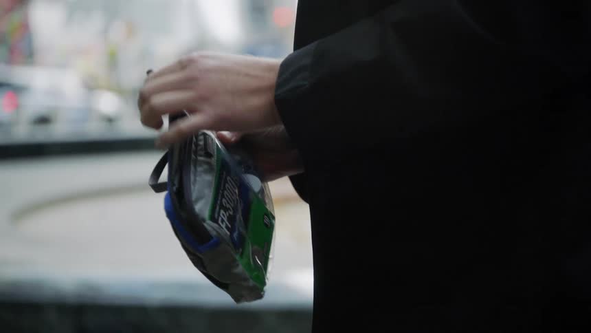 A man removes an object from a small zippered bag as he stands on a city sidewalk