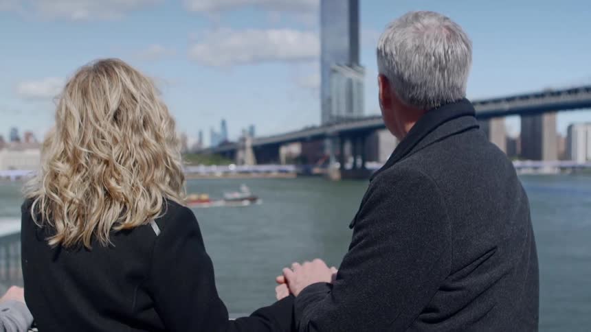 A man and woman hold hands standing next to a city river and bridge