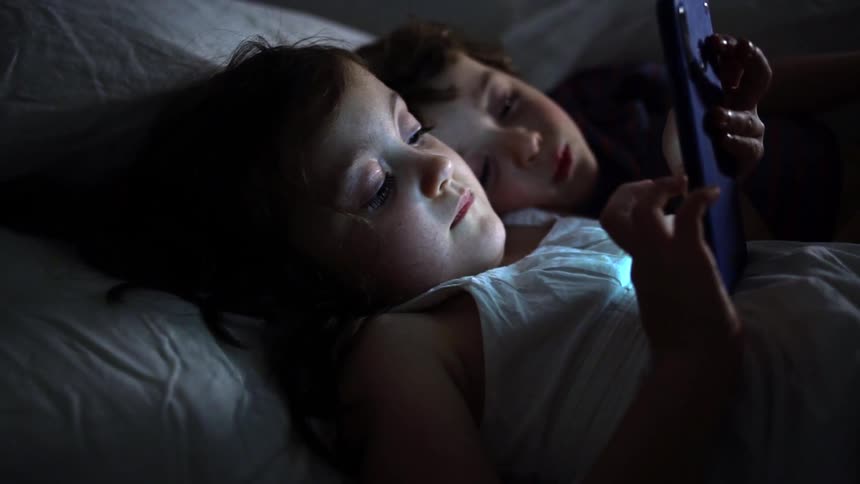 A brother and sister lie in bed and the girl plays on a cell phone