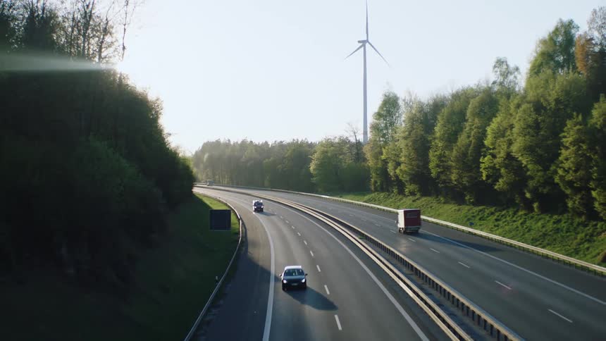 Vehicles drive along highway past woods and wind turbine