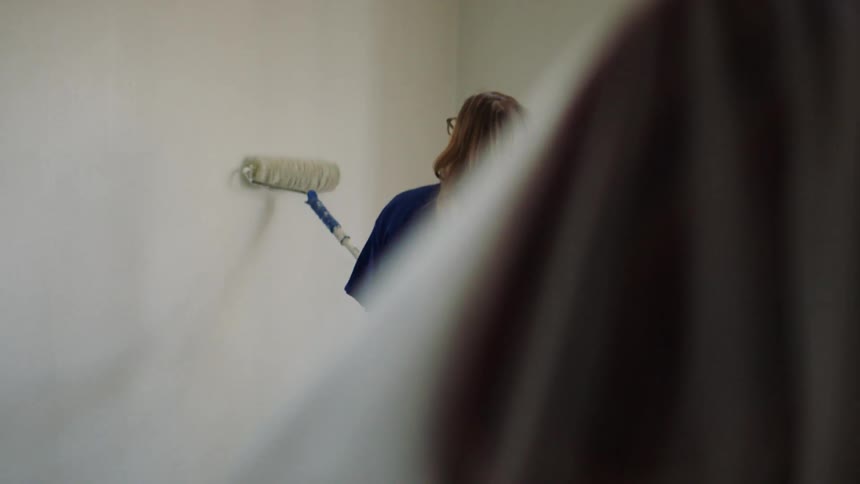 A woman uses a long paint roller to paint the top of the wall
