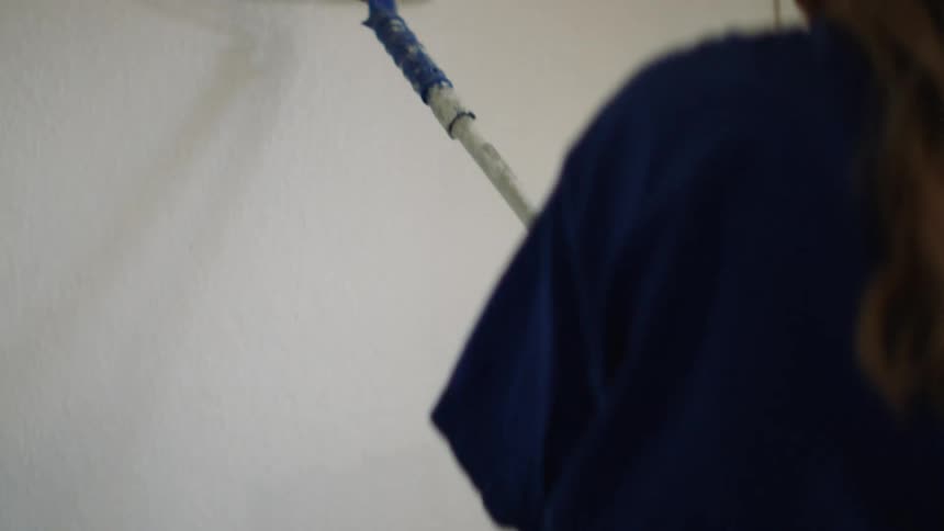 Female uses a paint roller with an extension pole to apply white paint to the wall in an up and down motion