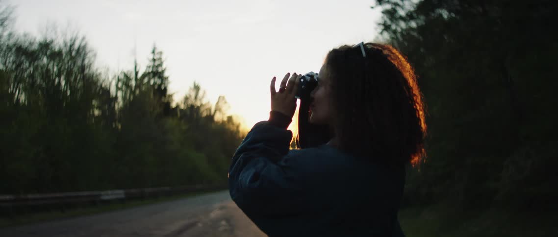 Female standing on the side of the country road surrounded by bushes and trees looks through her camera and adjusts the zoom lens as the sun sets in the horizon