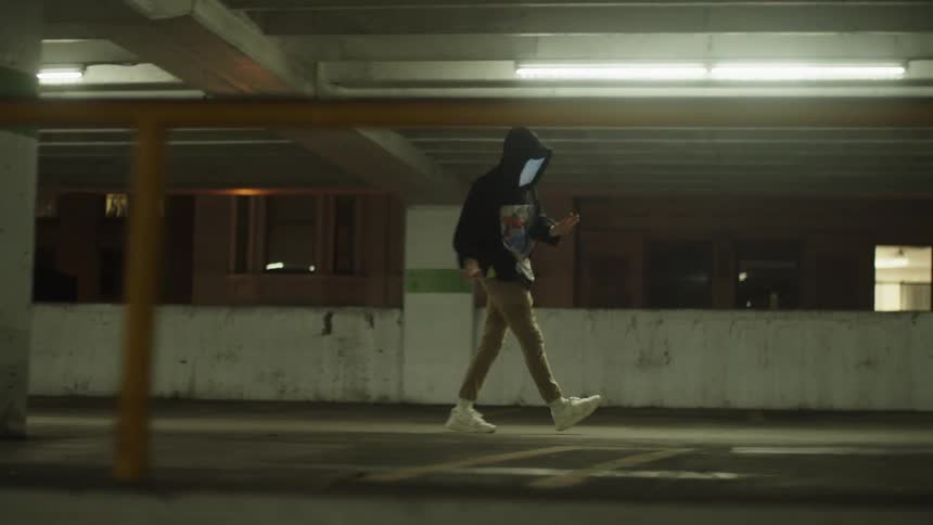 Hooded figure with a monitors for a face dances across an indoor car park