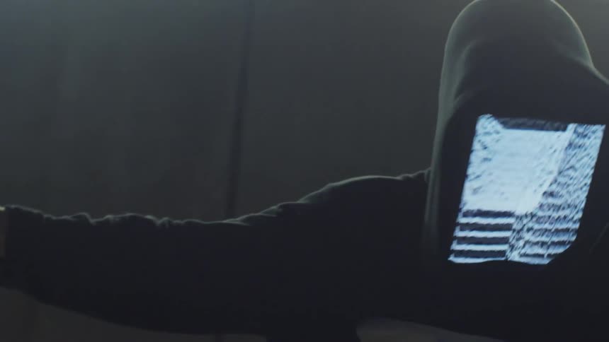 A white man with a monitor in his hood dances indoors