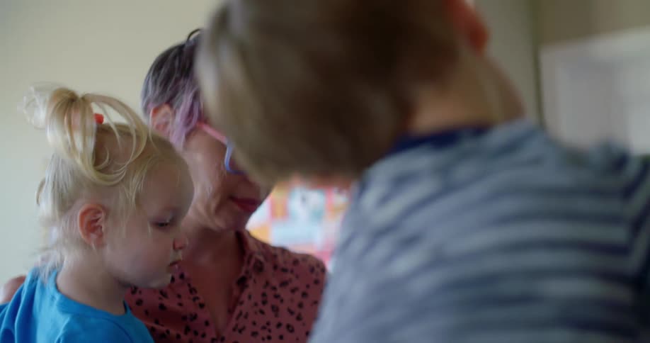 A mother indoors carries her daughter and speaks to her son