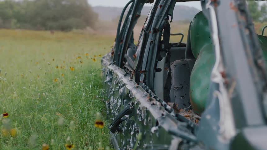 Abandoned vehicle covered in bullet holes in a field with overgrown weeds