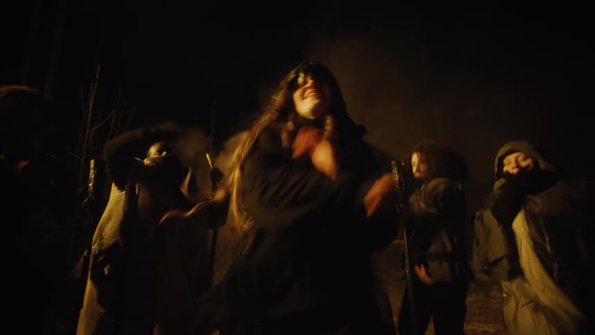 A group dressed in robes and rags dances at night with smoke billowing behind them