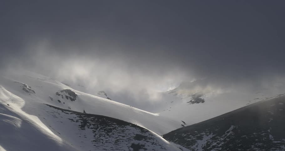 Aerial pulling up into dark clouds over snowy mountains at sunset