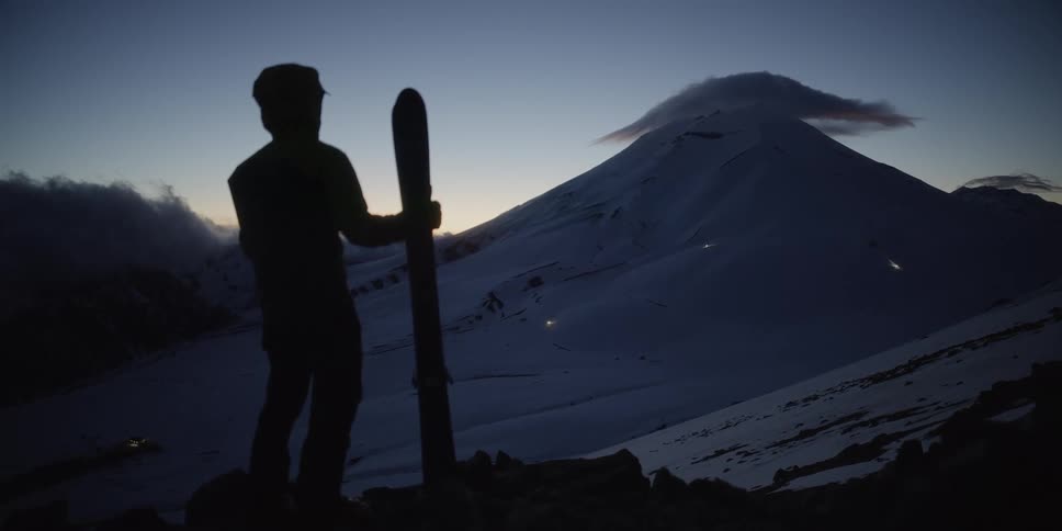 A man stands holding his skis as he looks towards at a snow covered mountain