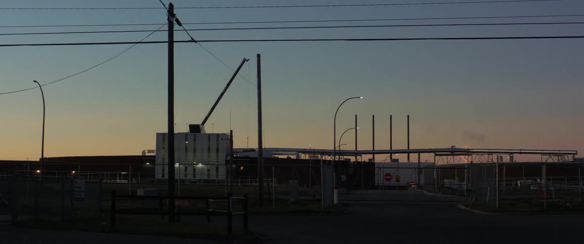 Cars travel on a road past a refinery with flames and smoke on the horizon at sunset