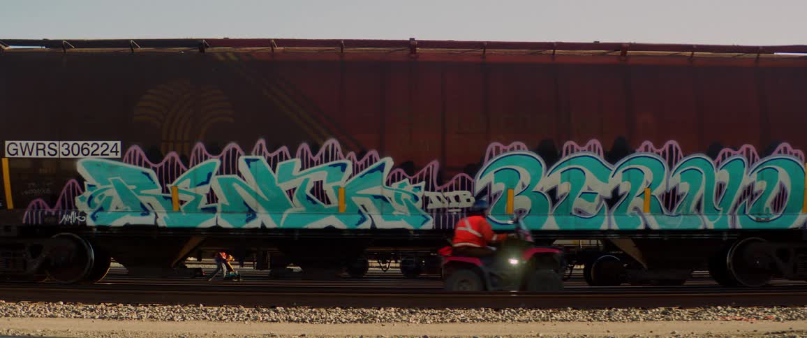 A parked train with people walking on one side and a man riding a 4 wheeler by on the other side