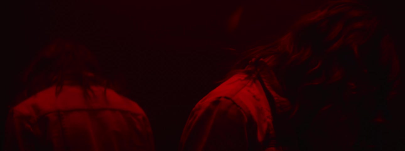 A man stands with his back to a mirror in the dark with his head down