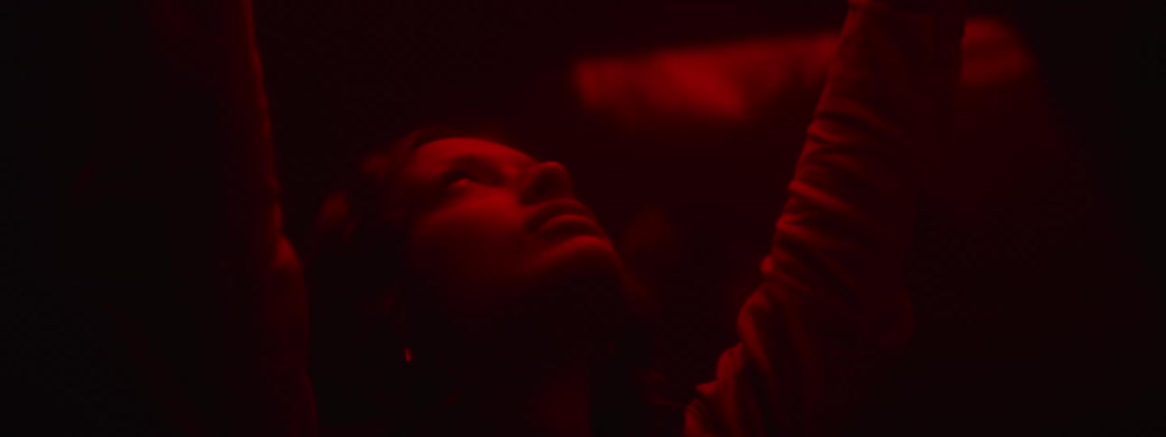 A woman stands with her arms stretched and reaching up and her head tilted back while a reddish light glows