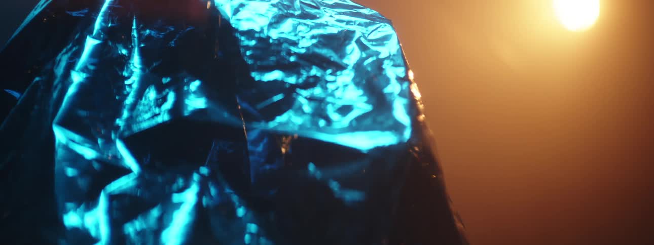 An object covered in a plastic trash bag in a dark misty room illuminated by a yellow spotlight