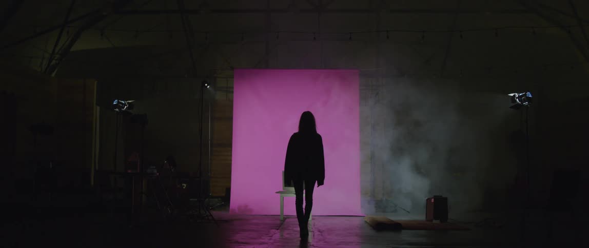 A female model wears a coat and walks across a dark stage with purple light
