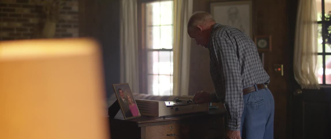 An elderly man walks across a living room and fiddles with a record player