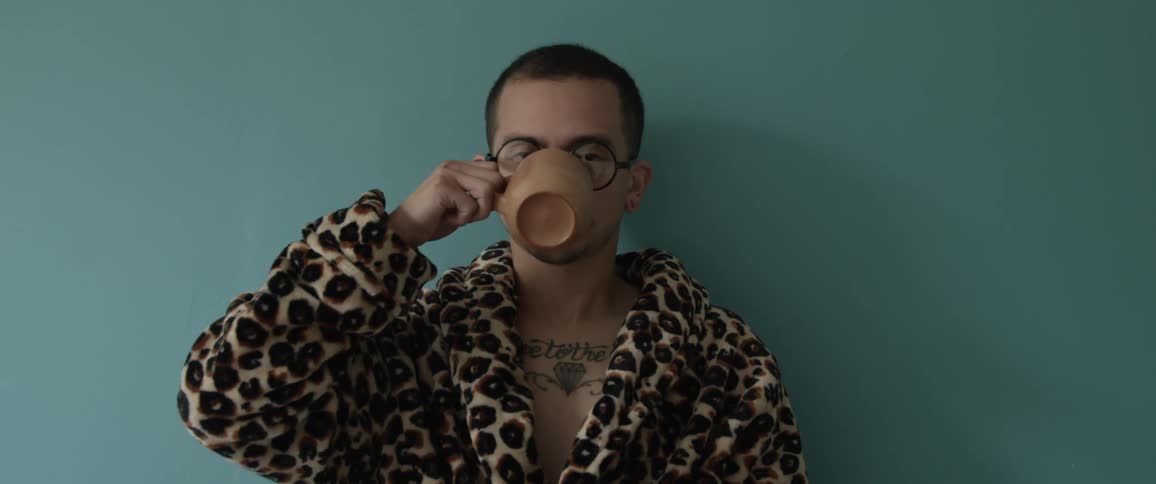 A white man drinks from a mug while wearing a robe in bed