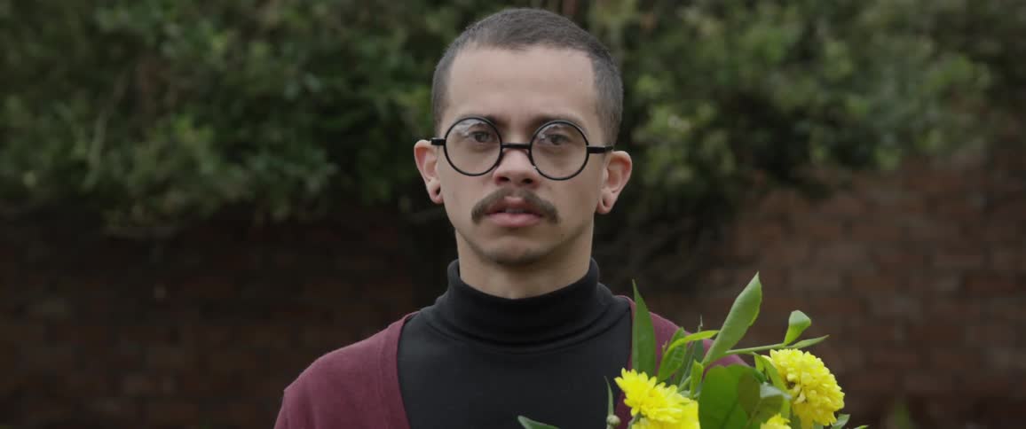 A white man holding flowers stands outdoors while taking a hit from an inhaler