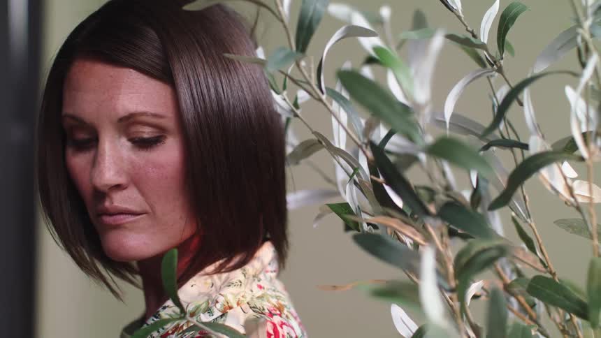 A woman stands by a plant and appears to be thinking about something