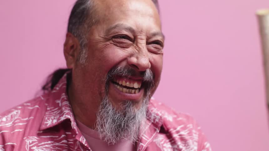 A man with a goatee and dressed in a pink shirt laughs and smiles